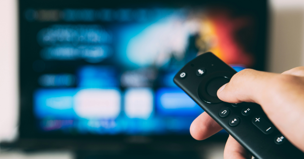 Mão segurando um controle remoto preto apontado para uma televisão ao fundo, que exibe uma interface de streaming com cores azuladas e alguns ícones visíveis.