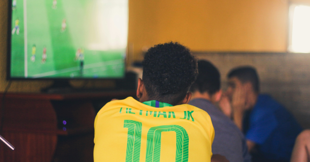 Pessoa vestindo uma camisa amarela da seleção brasileira com o nome 'Neymar Jr' e o número 10 estampados nas costas assiste a um jogo de futebol na TV.