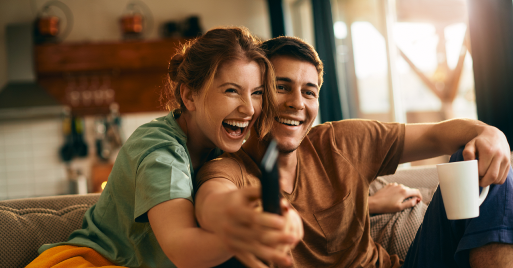 Casal sorridente sentado no sofá, disputando o controle remoto enquanto assiste à TV, representando a diversão e o entretenimento acessível proporcionados pelas vantagens do IPTV.